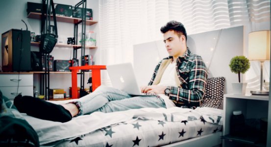 Man In Green Blue And Black Plaid Sports Shirt Sitting On Bed Using Silver Macbook photo