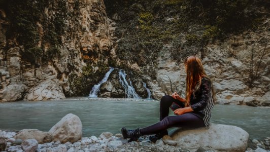 Woman With Brown Hair In Black Jacket Sitting On Rock Near Body Of Water photo