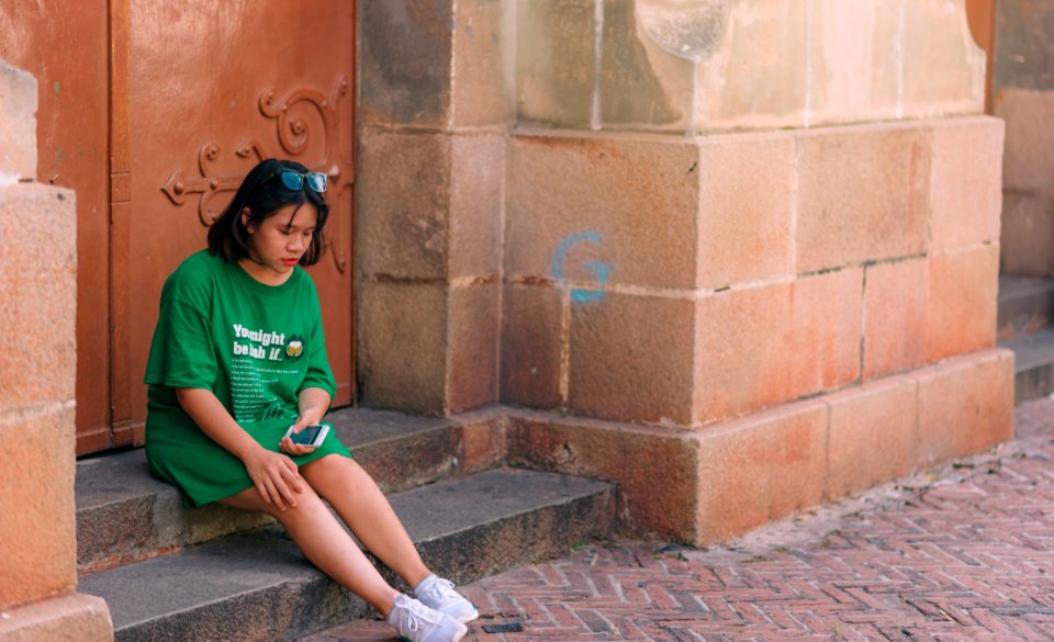 Woman Wearing Green Shirt Sitting Near Brown Gate Holding Smartphone