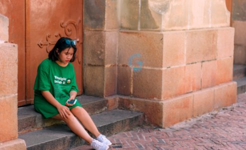Woman Wearing Green Shirt Sitting Near Brown Gate Holding Smartphone photo