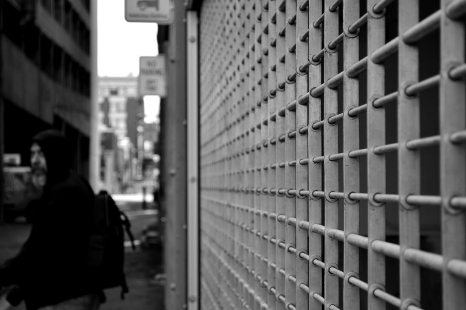 Grayscale Photo Of Man In Hoodie In Front Of White Window Grill