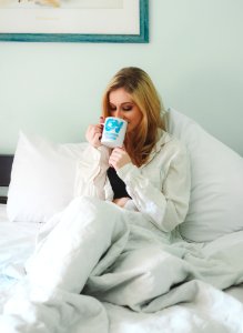 Woman Wearing White Dress Shirt Sitting On Bed While Drinking Through White Ceramic Mug photo