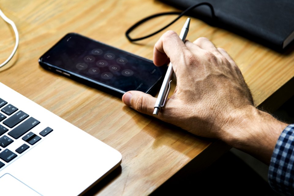 Person Holding Gray Retractable Pen And Black Iphone On Brown Wooden Table