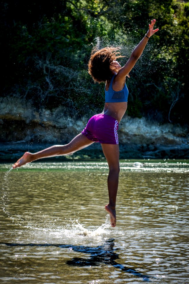 Woman In Blue Sports Bra And Purple Shorts Leaping Above Body Of Water