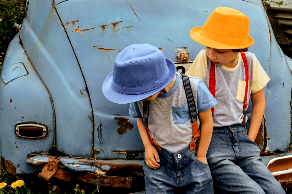 Two Boy In Grey Shirts And Blue Overall Pants Sitting On Blue Car Bumper