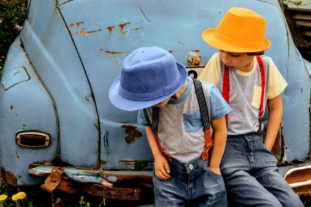 Two Boy In Grey Shirts And Blue Overall Pants Sitting On Blue Car Bumper