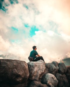 Man In Green Shirt And Black Pants Sitting On Top Of Rock Cliff Under White Clouds photo