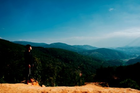 Man Stands On Cliff With Green Mountains On Background