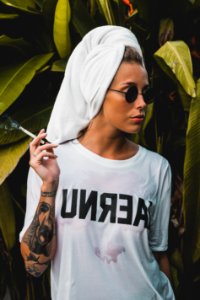 Woman Wearing White Crew-neck Shirt And Black Framed Sunglasses With White Bath Towel On Her Head Holding Cigarette Stick
