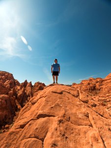 Man Wearing Blue Shirt And Black Shorts Standing On Top Of Brown Rock Formations Under Clear Sky