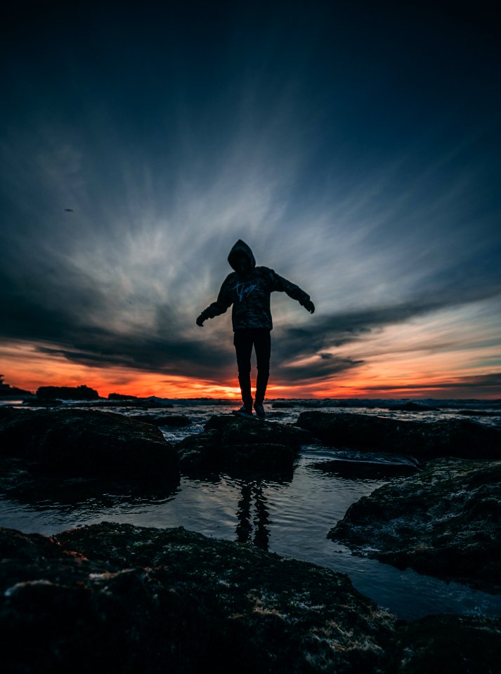 Person Wearing Hoodie Standing On Rock Surrounded By Body Of Water photo