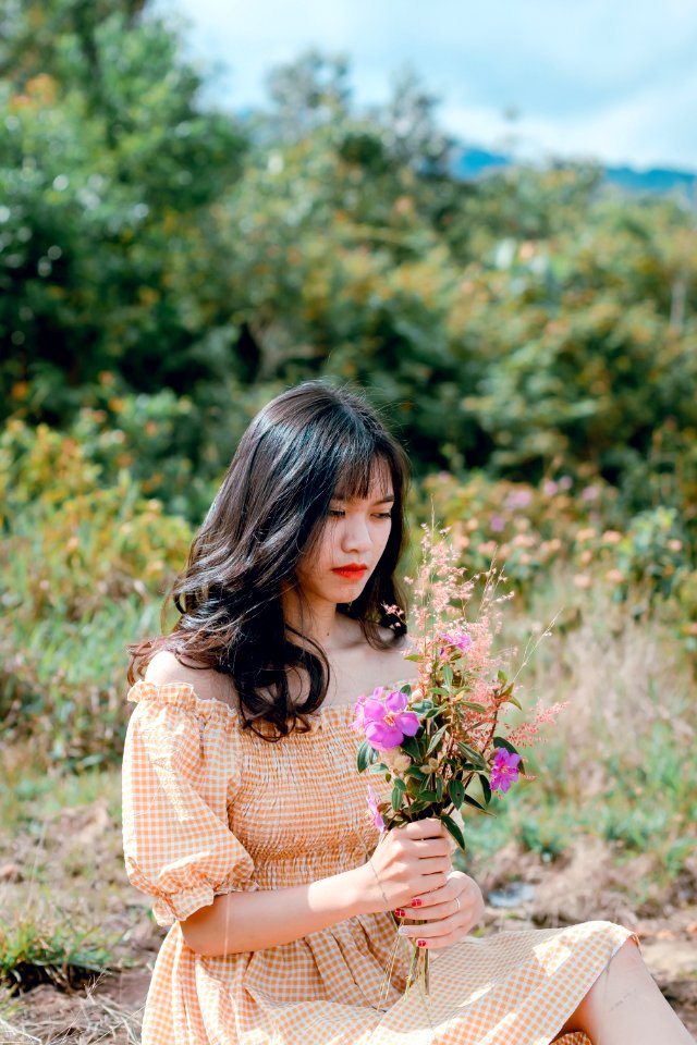 Shallow Focus Photography Of Woman Wearing Orange Checked Dress Holding Purple Flowers