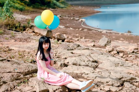 Woman In Pink Thick Strap Dress Holding A Yellow And Blue Balloons While Sitting On Rock Terrain photo
