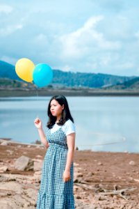 Woman Wearing Blue And White Plaid Dress Holding Blue And Yellow Balloons Near Calm Body Of Water At Daytime