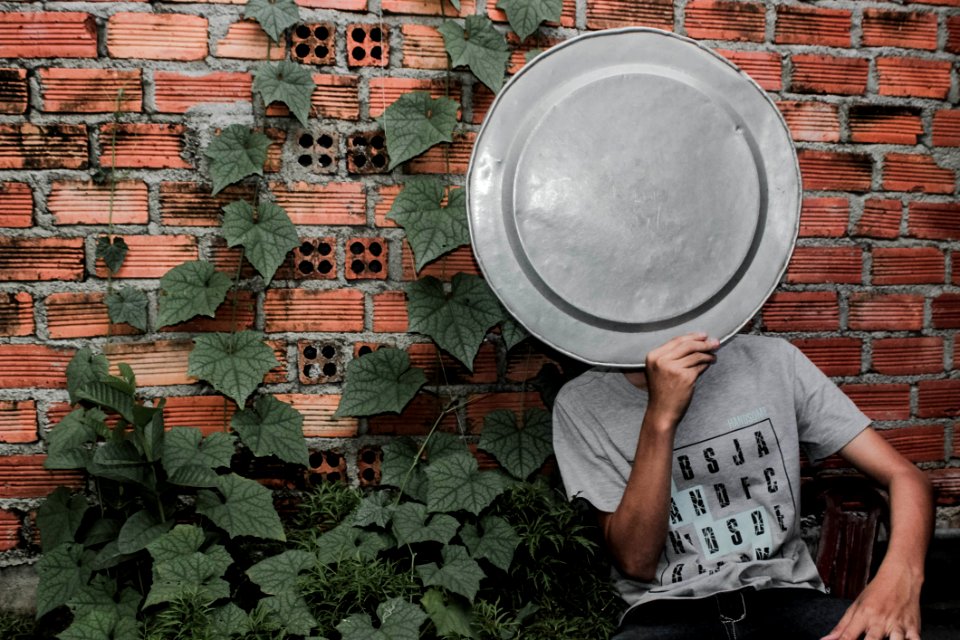 Person Wearing Gray Crew-neck T-shirt Holding A Tray On The Face Next To A Brick Wall