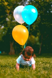 Baby Wearing White T-shirt Holding Three Yellow Blue And White Balloons On Green Grass Near Woods photo