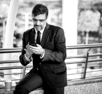 Greyscale Photo Of Man Wearing Suit Jacket And Eyeglasses Holding Smartphone photo