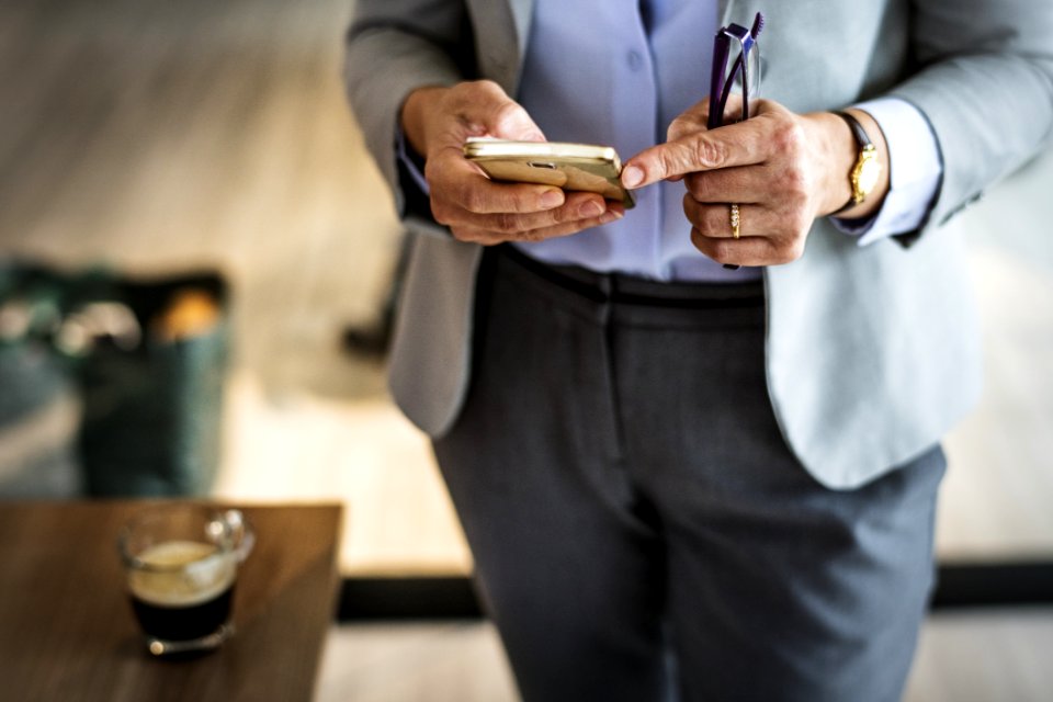 Person Wearing Grey Suit Jacket And Black Bottoms Standing Near Brown Wooden Table Holding Smartphone