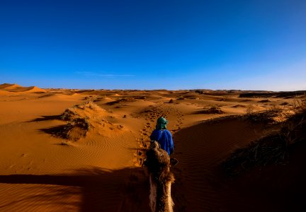 Man Wearing Blue Jacket Riding Camel Walking On Desert photo