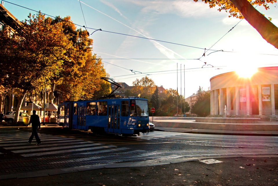 Person Wearing Black Walking On Pedestrian Lane Near Blue Tram Train During Sunset