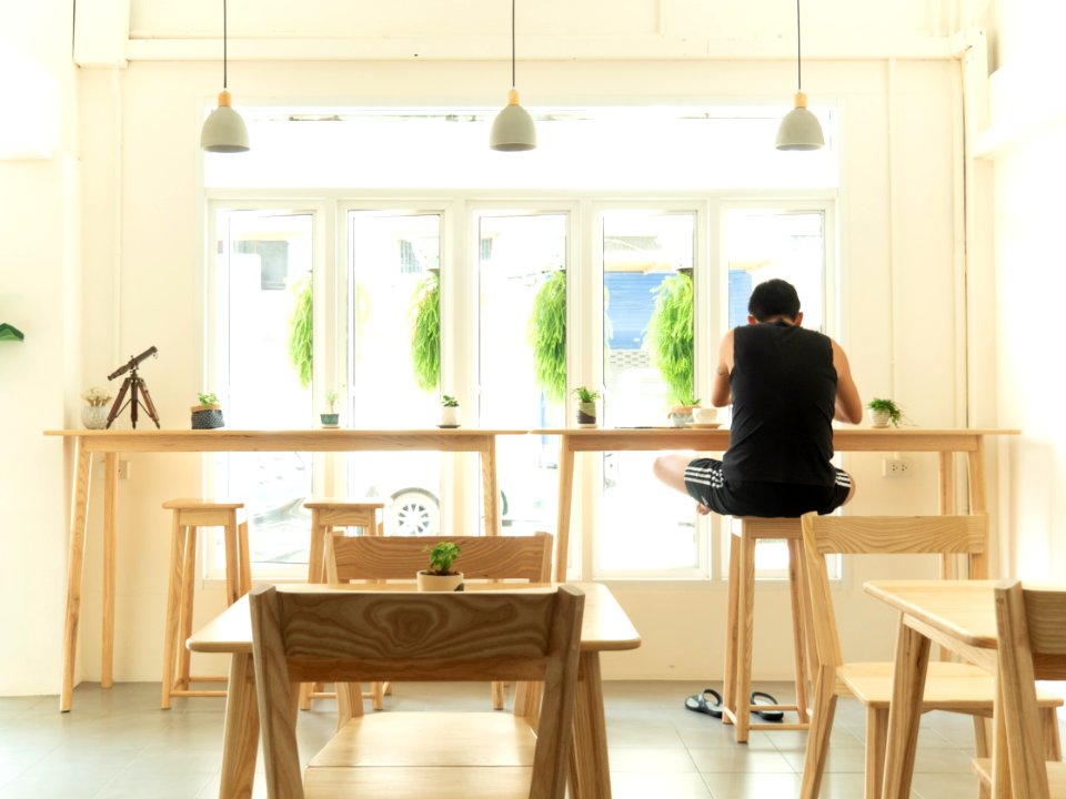 Man In Black Sleeveless Shirt Sitting On Wooden High Chair Beside Wooden Tables Near Window