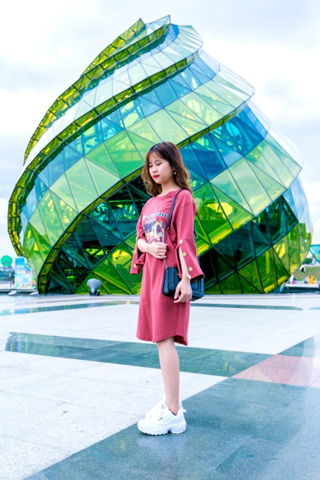 Girl Wearing Red Long-sleeved Dress And White Low-top Shoes Standing Near Green Glass Structure