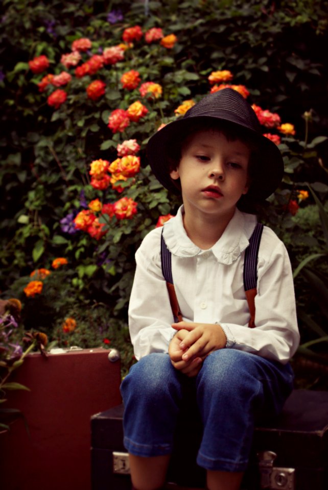 Boy Wearing Black Hat Sitting On Case Near Flowers