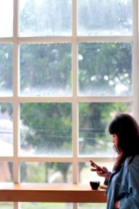 Woman Wearing Blue Denim Jacket Holding Smartphone Standing Beside Clear Glass Window photo