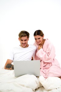 Man In White Crew-neck Shirt Sitting On Bed Beside Woman In Pink Bathrobe Looking At Laptop Computer photo