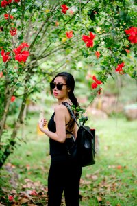 Woman Looking Over Her Shoulder Wearing Black Sleeveless Top And Black Jeans Under A Red Hibiscus Plant At Daytime