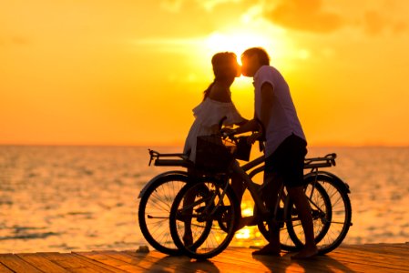 Photography Of Man Wearing White T-shirt Kissing A Woman While Holding Bicycle On River Dock During Sunset photo