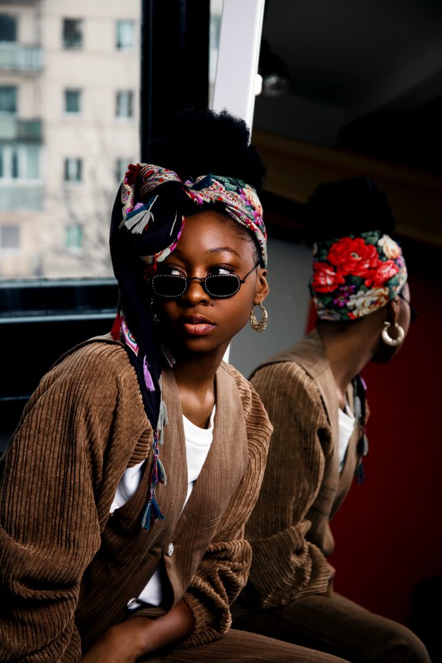 Woman Wearing White Shirt And Brown Coat With Sunglasses And Floral Turban Taking Selfie With Mirror Beside Her