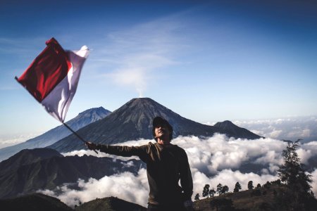 Man Wearing Black Crew-neck Sweater Holding White And Red Flag Standing Near Mountain Under Blue And White Sky photo