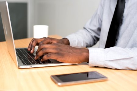 Person Wearing White Dress Shirt And Black Necktie Using Macbook Air On Beige Wooden Table