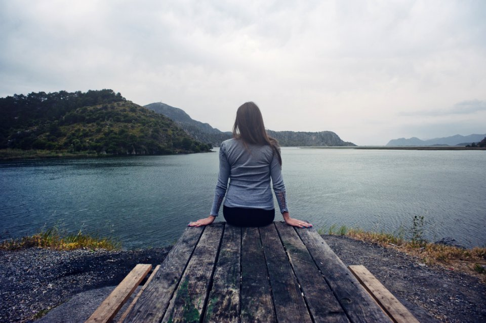 Woman Wearing Gray Long-sleeved Shirt And Black Black Bottoms Outfit Sitting On Gray Wooden Picnic Table Facing Towards Calm Body