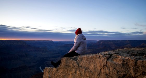 Person Wearing Gray Hooded Jacket And Black Pants Sitting On Mountain Cliff During Sunset photo