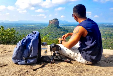 Man Sitting On Top Of Gray Cliff Mountain Beside Backpack Water Bottle And Camera photo