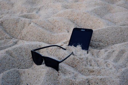 Black Wayfarer-style Sunglasses Beside Black Asus Android Smartphone On Brown Sand