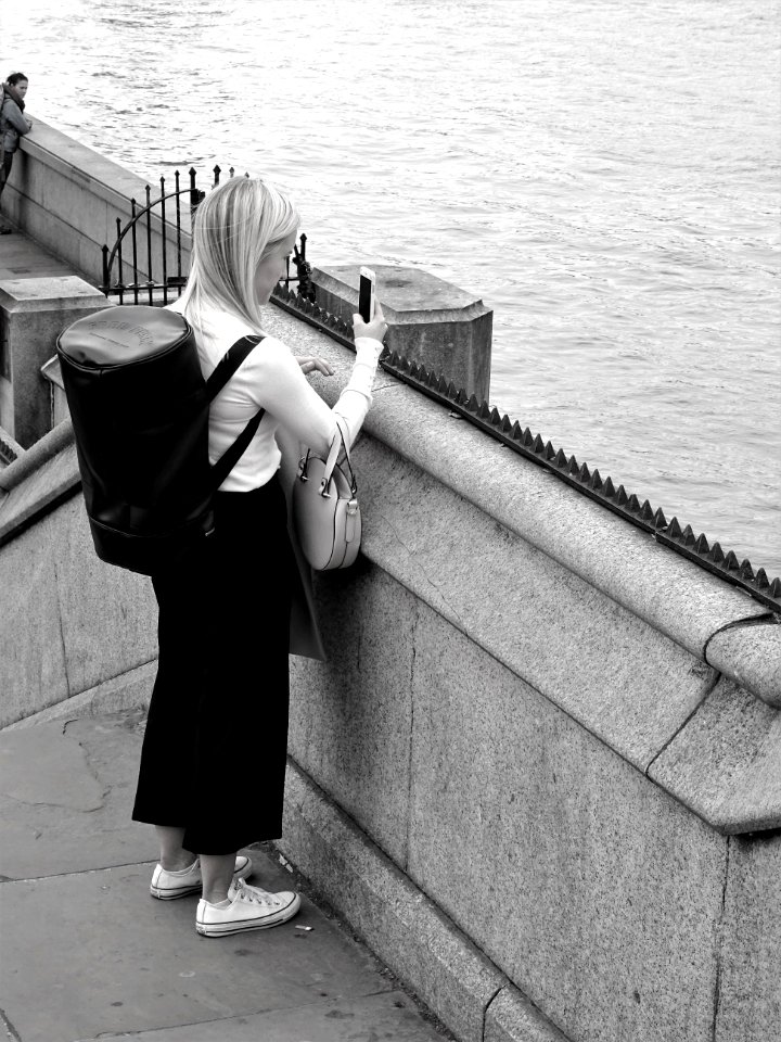 Black And White Photo Of Woman Holding Smartphone While Standing Near Calm Water photo