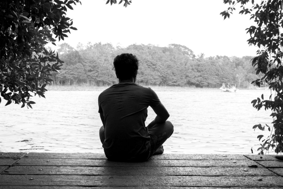 Gray Scale Photography Of Man Sitting On Brown Wooden Floor Beside Body Of Water