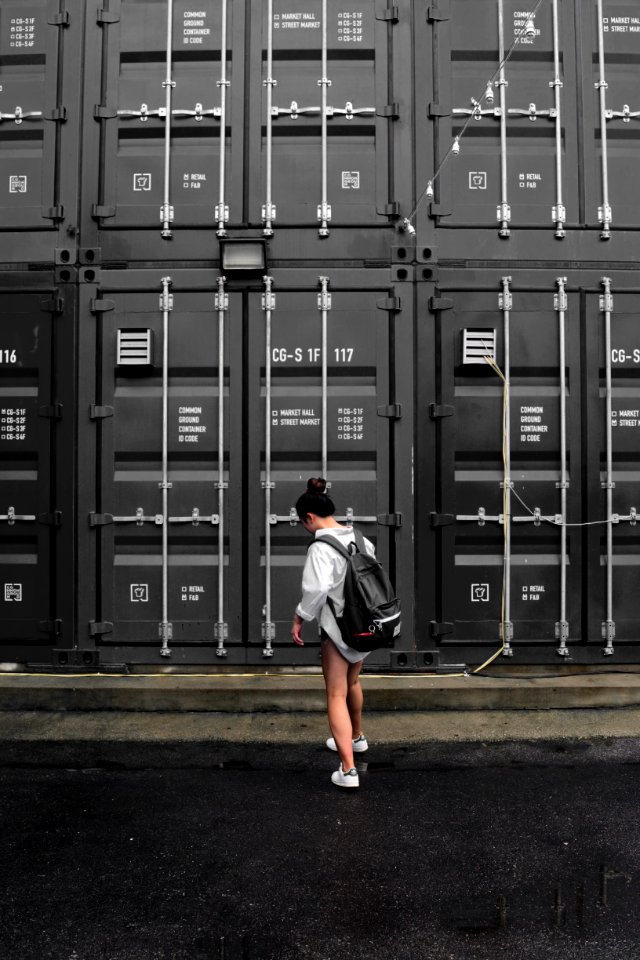 Photography Of Woman In Gray Long Sleeve Shirt Carrying Black Backpack Standing In Front Of Black Intermodal Container