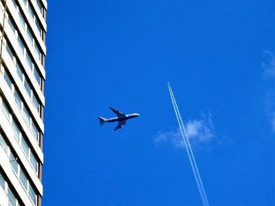 Low-angle Photography Of White Plane On Mid-air Near White Concrete Building And White Contrail photo