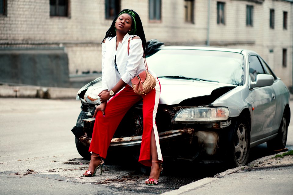 Woman In White Open Cardigan And Red And White Pants Sitting On Damage White Car