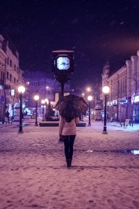 Person In White Long-sleeved Top And Black Pants Under Black Umbrella Standing Near Street Clock