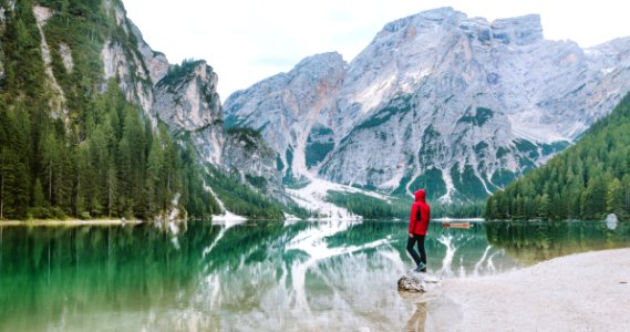 Man Wearing Red Hoodie Standing Near Body Of Water With View Of Mountains