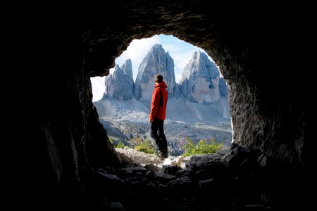 Man In Red Jacket Standing Outside Of Cave In Front Of Three Mountains photo