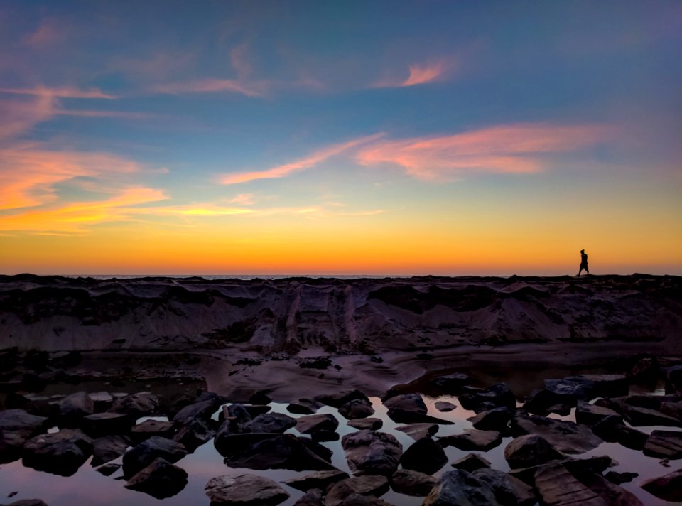 Silhouette Photo Of Man Walking On Hill Near Body Of Water And Rock Formations