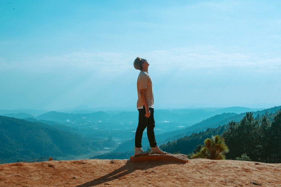 Man Wearing Beige Shirt And Black Pants Looking Up With Blue Sky And Mountains In The Background