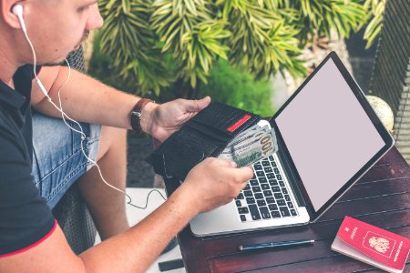 Man In Black T-shirt And Blue Denim Shorts Sitting While Pulling Out 100 Us Dollar Banknotes From Black Wallet