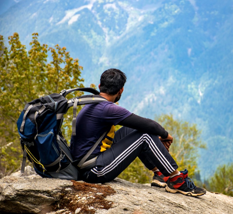 Man In Purple T-shirt With Blue Backpack Sitting On Gray Boulder